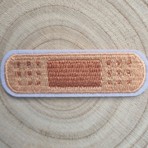 May include: Embroidered patch in the shape of a plaster. The patch features a light brown centre with a darker brown rectangular pad and small dotted details. The patch is outlined in a light blue colour.