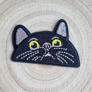May include: A black cat patch with embroidered details. The cat has yellow eyes, a pink mouth, and white whiskers. The patch is on a light brown, wooden surface.