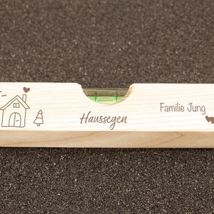 Wasserwaage aus Holz mit Gravur. In der Mitte steht Haussegen, links ist ein kleines Haus mit Herz und Baum zu sehen, Rechts steht ein Familienname mit zwei kleinen Herzchen