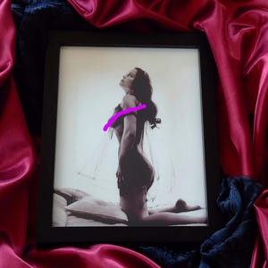 May include: A black framed photograph of a woman in a sheer garment, kneeling on a white pillow. The woman is looking up and her hair is long and dark. The background is a light gray.