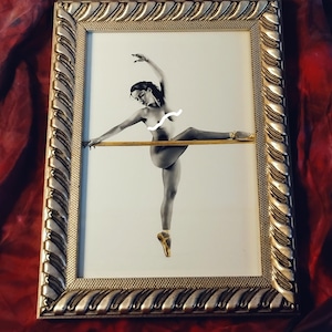 May include: Black and white framed print of a ballerina in a dance pose. The ballerina is wearing pointe shoes and is posed with one leg extended. The frame is gold with a textured design. The background is a red fabric.