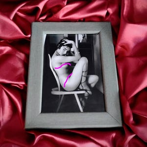 May include: A framed black and white photograph of a person seated on a chair, with arms crossed. The photograph is in a gray wooden frame, set against a backdrop of red fabric.