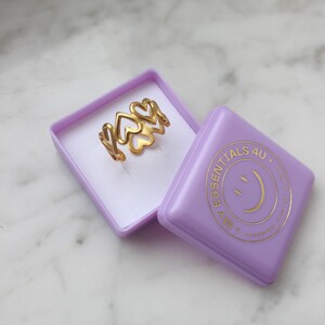 Peut inclure: Une bague dor&eacute;e avec un motif de c&oelig;ur, pr&eacute;sent&eacute;e dans une bo&icirc;te lavande. La bague pr&eacute;sente plusieurs formes de c&oelig;ur. La bo&icirc;te porte le texte "ESSENTIALS 4U" et un logo de smiley.