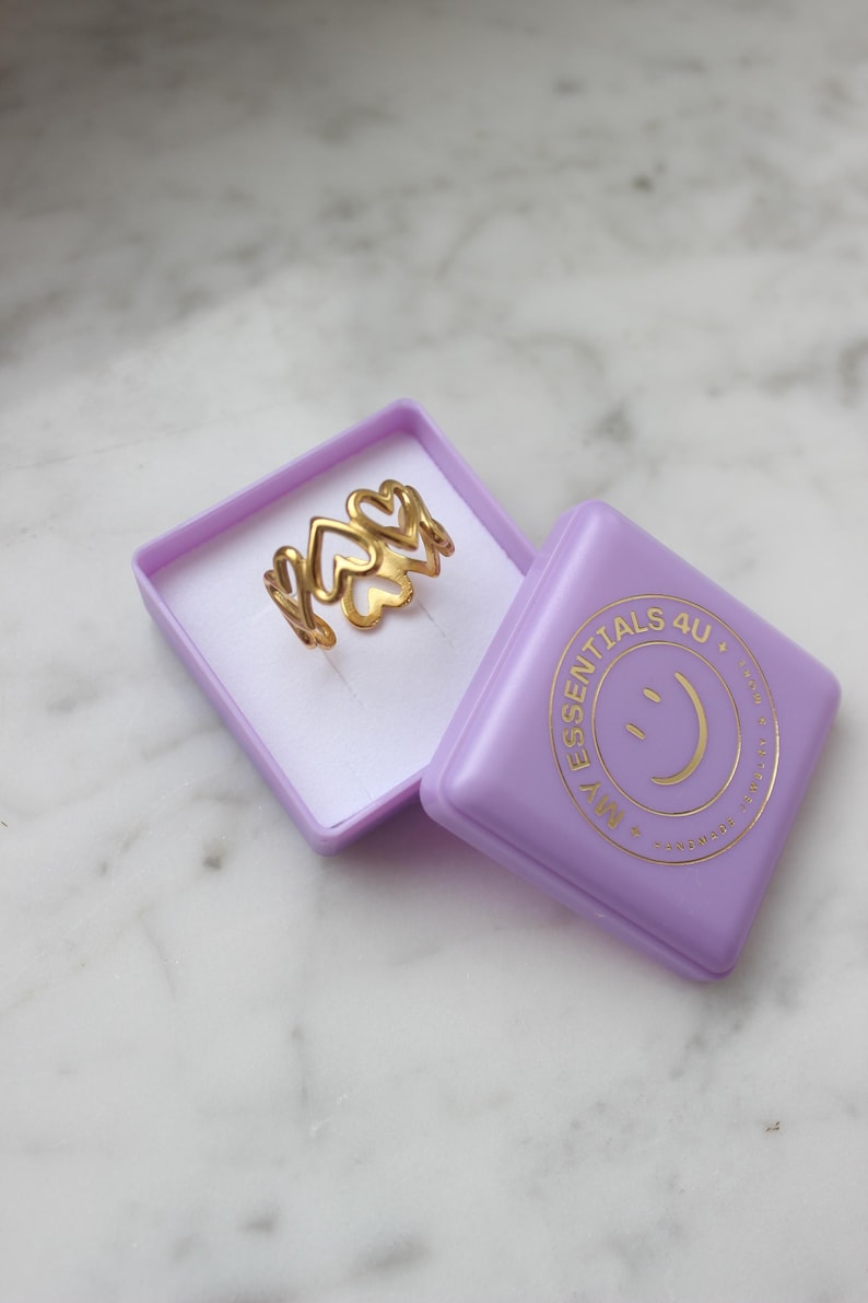 Peut inclure: Une bague dor&eacute;e avec un motif de c&oelig;ur, pr&eacute;sent&eacute;e dans une bo&icirc;te lavande. La bague pr&eacute;sente plusieurs formes de c&oelig;ur. La bo&icirc;te porte le texte "ESSENTIALS 4U" et un logo de smiley.