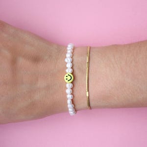 Könnte beinhalten: Zwei Armbänder an einem Handgelenk vor rosa Hintergrund. Ein Armband hat kleine weiße Perlen und eine gelbe Smiley-Gesicht-Perle. Das andere ist ein dünnes goldenes Kettenarmband.