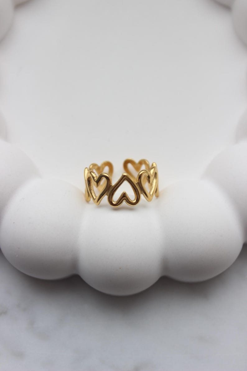 Peut inclure: Une bague dor&eacute;e orn&eacute;e d'une s&eacute;rie de d&eacute;coupes en forme de c&oelig;ur. La bague &agrave; design ouvert est pos&eacute;e sur une surface blanche en forme de nuage. La bague est un bijou.