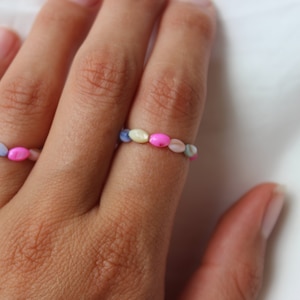 Könnte beinhalten: Zwei bunte Perlenringe an einer Hand. Die Ringe bestehen aus kleinen, ovalen Perlen in den Farben Rosa, Blau und Weiß.