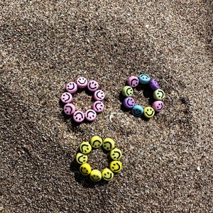 Könnte beinhalten: Drei bunte Armbänder aus kleinen Smiley-Perlen. Die Armbänder sind rosa, lila und gelb. Die Armbänder sind in einem Kreis auf einer sandigen Oberfläche angeordnet.
