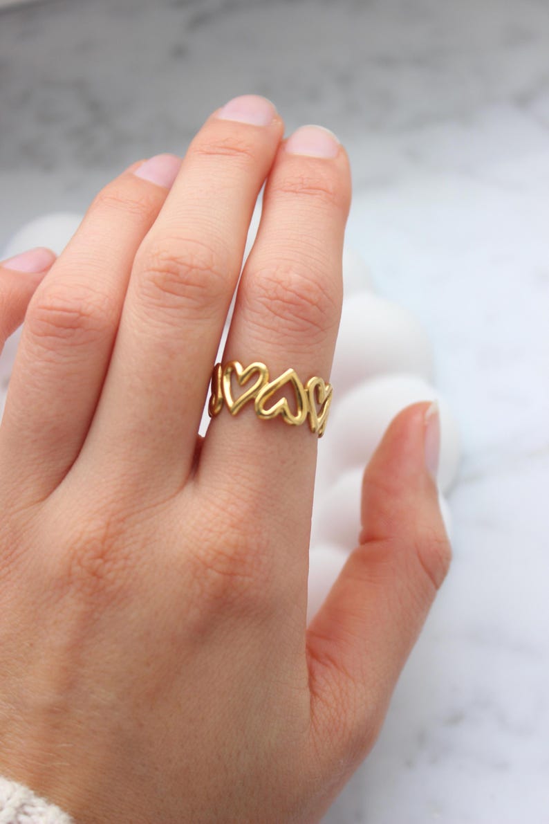 Peut inclure: Une bague dor&eacute;e orn&eacute;e d'une s&eacute;rie de c&oelig;urs ajour&eacute;s. La bague est port&eacute;e au doigt, mettant en valeur son design d&eacute;licat. La teinte chaude de la bague contraste avec le teint de la peau, soulignant l'attrait esth&eacute;tique du bijou.