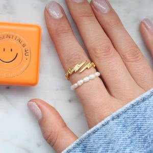 Op de afbeelding: Een gouden ring met een bliksemontwerp en een parelring aan dezelfde vinger. De ring zit aan een hand met witte glitternagellak.
