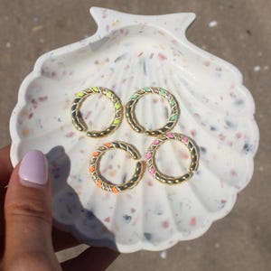Könnte beinhalten: Vier goldfarbene Creolen mit farbigen Emaille-Akzenten in Neon-Gelb, Mintgrün, Orange und Pink. Die Ohrringe sind auf einer weißen, muschelförmigen Schale mit einem gesprenkelten Muster angeordnet. Die Ohrringe haben ein gedrehtes Seil-Design.