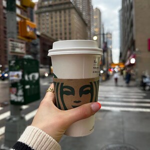 Peut inclure: Une tasse &agrave; caf&eacute; en papier blanche avec un couvercle et un manchon marron Starbucks est tenue par une personne. La personne porte un pull cr&egrave;me avec des rayures noires sur la manche. En arri&egrave;re-plan, une rue de la ville.