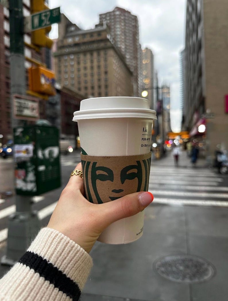 Peut inclure: Une tasse &agrave; caf&eacute; en papier blanche avec un couvercle et un manchon marron Starbucks est tenue par une personne. La personne porte un pull cr&egrave;me avec des rayures noires sur la manche. En arri&egrave;re-plan, une rue de la ville.