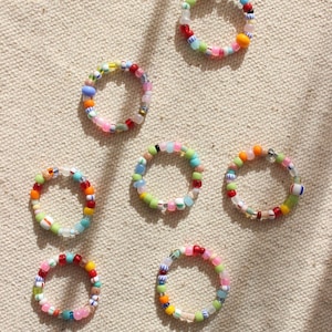 Puede incluir: Una colección de anillos de cuentas coloridas. Cada anillo presenta una combinación única de pequeñas cuentas redondas en varios colores, incluyendo rojo, amarillo, azul, verde y rosa. Los anillos están dispuestos sobre una superficie texturizada de tono neutro.