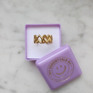 Peut inclure: Une bague dor&eacute;e avec un motif de c&oelig;urs, pr&eacute;sent&eacute;e dans une bo&icirc;te violette. La bague comporte plusieurs formes de c&oelig;urs. Une bo&icirc;te violette avec le texte "MY ESSENTIALS 4U" et un smiley est &agrave; c&ocirc;t&eacute;.
