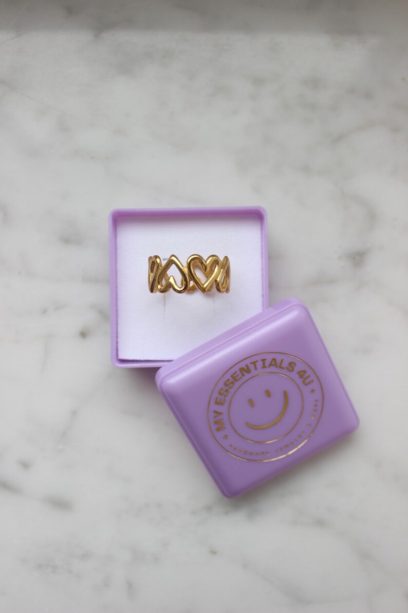 Peut inclure: Une bague dor&eacute;e avec un motif de c&oelig;urs, pr&eacute;sent&eacute;e dans une bo&icirc;te violette. La bague comporte plusieurs formes de c&oelig;urs. Une bo&icirc;te violette avec le texte "MY ESSENTIALS 4U" et un smiley est &agrave; c&ocirc;t&eacute;.