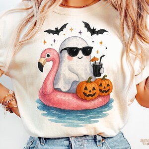 May include: Cream-colored t-shirt featuring a whimsical Halloween design. A cartoon ghost wearing sunglasses floats on a pink flamingo floatie in water. Two jack-o'-lanterns and bats complete the festive scene. The shirt has the text "Annia Digital Art".