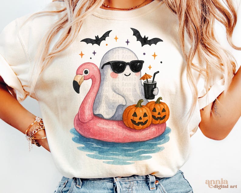 May include: Cream-colored t-shirt featuring a whimsical Halloween design. A cartoon ghost wearing sunglasses floats on a pink flamingo floatie in water. Two jack-o'-lanterns and bats complete the festive scene. The shirt has the text "Annia Digital Art".
