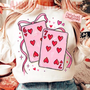 May include: White sweatshirt featuring a pink design of playing cards with hearts. The cards are adorned with red hearts and pink ribbons. The numbers 6 and 7 are visible on the cards. The words "Original Designer" are also visible.