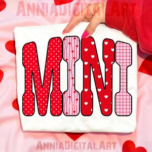 May include: A white t-shirt with the word "MINI" in a bold, decorative font. The letters are red and pink, featuring polka dots, stripes, and heart patterns. The background is a pink fabric with red heart shapes.