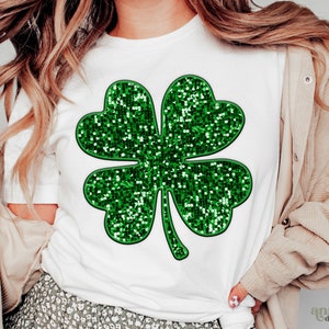 Puede incluir: Una camiseta blanca con un diseño de trébol de cuatro hojas con purpurina verde.
