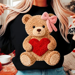 May include: Black long-sleeved sweatshirt featuring a plush teddy bear holding a red heart. The teddy bear has a pink bow and is centered on the front. The sweatshirt is a solid black color.