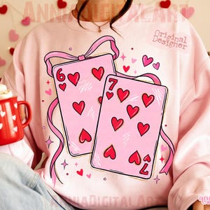 May include: A light pink sweatshirt featuring a graphic of two playing cards with red hearts. The cards are adorned with pink ribbons and the numbers 6 and 7. The sweatshirt has the text "Original Designer" and "annia digital art".