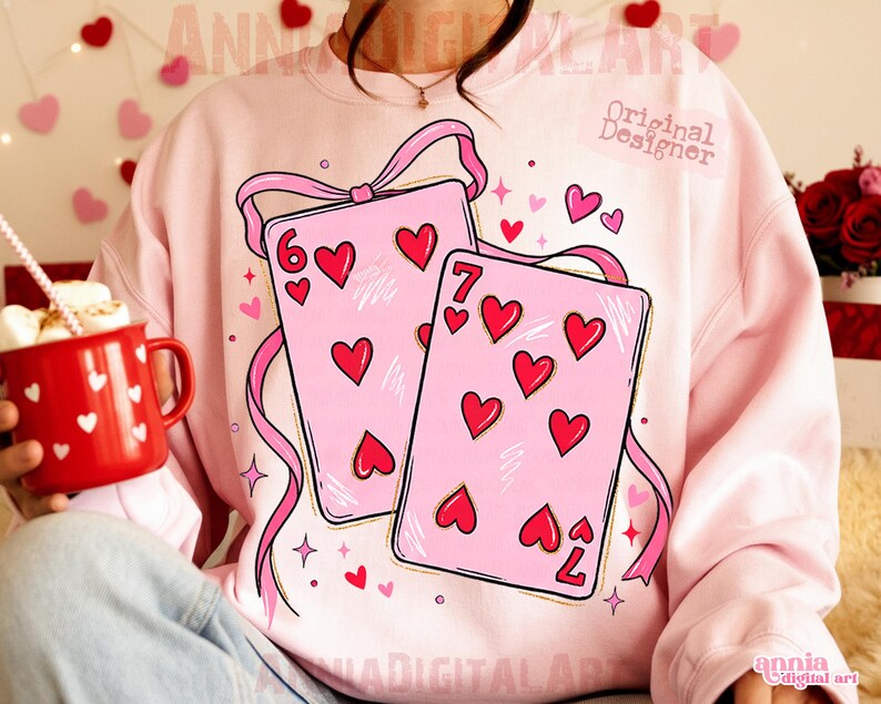 May include: A light pink sweatshirt featuring a graphic of two playing cards with red hearts. The cards are adorned with pink ribbons and the numbers 6 and 7. The sweatshirt has the text "Original Designer" and "annia digital art".