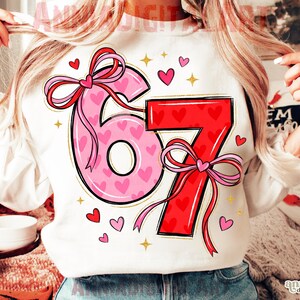 May include: White sweatshirt with the numbers "67" in pink and red, decorated with hearts and bows. The number "6" is pink with small pink hearts, and the number "7" is red with red hearts. Gold stars and small hearts are scattered around the numbers.