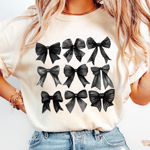 May include: A white t-shirt with a black and white graphic of nine different bows. The bows are all different shapes and sizes, and some have polka dots.