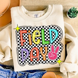 May include: A cream-colored sweatshirt with a checkered background and the words "Field Day" in rainbow colors. A pink peace sign is next to the text.