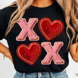 Op de afbeelding: Zwart T-shirt met roze en rode pluche letters en harten. Het ontwerp toont de letters "X" en harten in roze en rood, omlijnd met goud. Het shirt heeft een casual stijl.