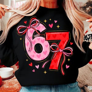 May include: Black sweatshirt featuring the numbers "67" in a pink and red design. The number "6" is pink and filled with small hearts, while the "7" is red. Both numbers have bow accents. Small hearts and stars surround the numbers.