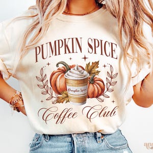May include: A white t-shirt with the text "Pumpkin Spice Coffee Club" in brown lettering. The design features a cup of pumpkin spice coffee with whipped cream and two pumpkins with fall leaves.