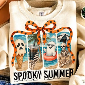 May include: Cream-colored sweatshirt with a "Spooky Summer" graphic. The graphic features a ghost ice cream cone, a skeleton holding a drink, and a ghost in a floatie on a beach scene. An orange bow with black polka dots frames the design.