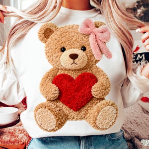 May include: White sweatshirt featuring a plush teddy bear design. The teddy bear is brown with a red heart and a pink bow. The sweatshirt is a cozy and cute design, perfect for Valentine's Day or everyday wear.