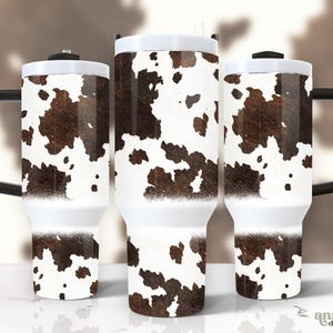May include: Three white travel mugs with black handles and lids. The mugs feature a cowhide pattern in shades of brown and white. The design is printed on the body of the mugs, with a white base and a black handle.