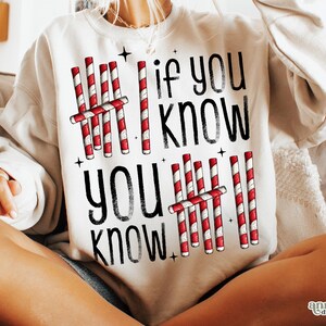 May include: Cream-colored sweatshirt with the text "If You Know, You Know" in black. The design includes red and white candy cane graphics and small black star accents, creating a festive holiday theme.