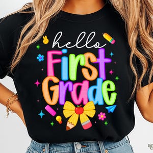 May include: Black t-shirt featuring the phrase "hello First Grade" in vibrant, bubble-style lettering. The design incorporates a yellow bow with a red apple, a pencil, and other school-related illustrations. The shirt is a casual wear.