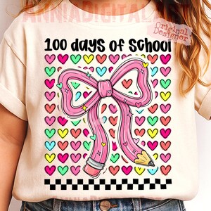 May include: Cream-colored t-shirt with the text "100 days of School." The shirt features a pink pencil shaped like a bow, surrounded by colorful hearts. A black and white checkered pattern is at the bottom.