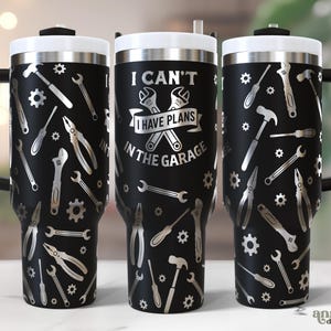 May include: Three black travel mugs with silver-colored tools and gears. The central mug has the text "I CAN'T I HAVE PLANS IN THE GARAGE" with crossed wrenches. Each mug has a black handle and a white rim.