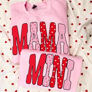 May include: Two light pink sweatshirts. The top sweatshirt says "MAMA" in red letters with white polka dots, stripes, and a grid pattern. The bottom sweatshirt says "MINI" in a similar style. The background is white fabric with red hearts.