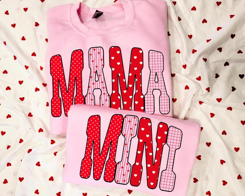 May include: Two light pink sweatshirts. The top sweatshirt says "MAMA" in red letters with white polka dots, stripes, and a grid pattern. The bottom sweatshirt says "MINI" in a similar style. The background is white fabric with red hearts.