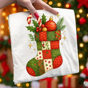 May include: A white t-shirt featuring a Christmas stocking design. The stocking is patchwork, with red and green squares, stars, and festive embellishments. A candy cane, ornaments, and holly complete the holiday theme.