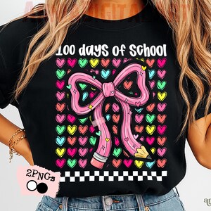 May include: Black t-shirt with the text "100 days of school." The design features a pink pencil shaped like a bow, surrounded by colorful hearts. A black and white checkered pattern is at the bottom.