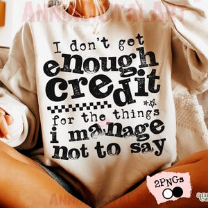 May include: Cream-colored sweatshirt with the text "I don't get enough credit for the things I manage not to say" in black. The design includes a star and a checkered pattern. The sweatshirt is a casual style.