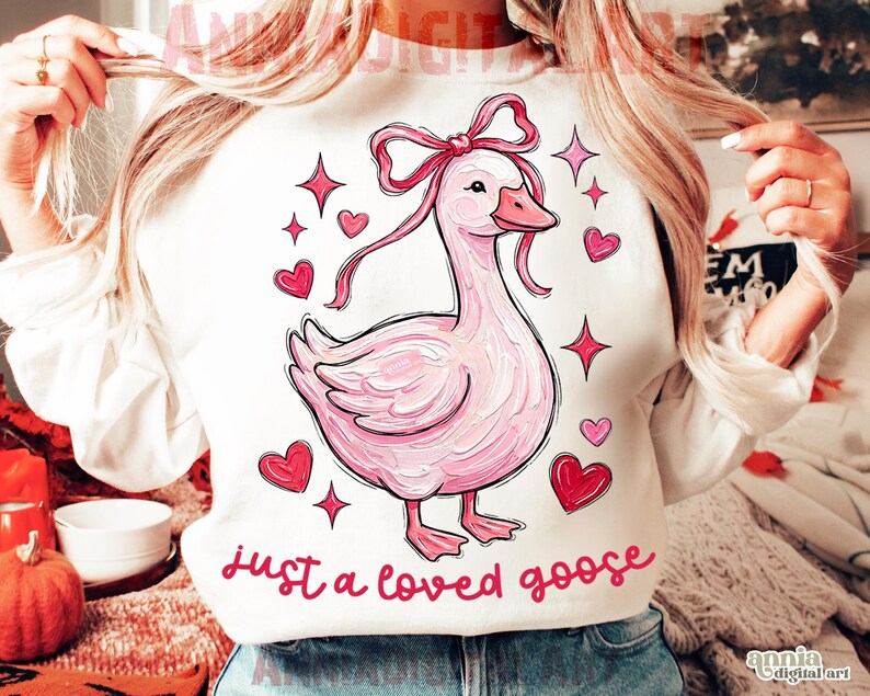Pu&ograve; includere: Felpa bianca con un'illustrazione di un'oca rosa, fiocco, cuori e stelle. Il testo "just a loved goose" &egrave; scritto in rosa. Un design affascinante e adorabile.