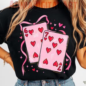 May include: Black t-shirt featuring a design of two pink playing cards with red hearts, the six of hearts and the seven of hearts. Pink ribbons and hearts surround the cards. The shirt has rolled-up sleeves.