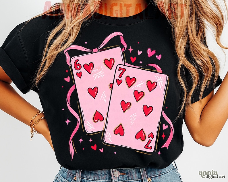 May include: Black t-shirt featuring a design of two pink playing cards with red hearts, the six of hearts and the seven of hearts. Pink ribbons and hearts surround the cards. The shirt has rolled-up sleeves.