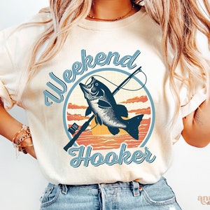 Pode incluir: Uma camiseta branca com um gráfico de estilo vintage de um peixe em um anzol de pesca. O texto "Weekend Hooker" está escrito em uma fonte de letra azul.